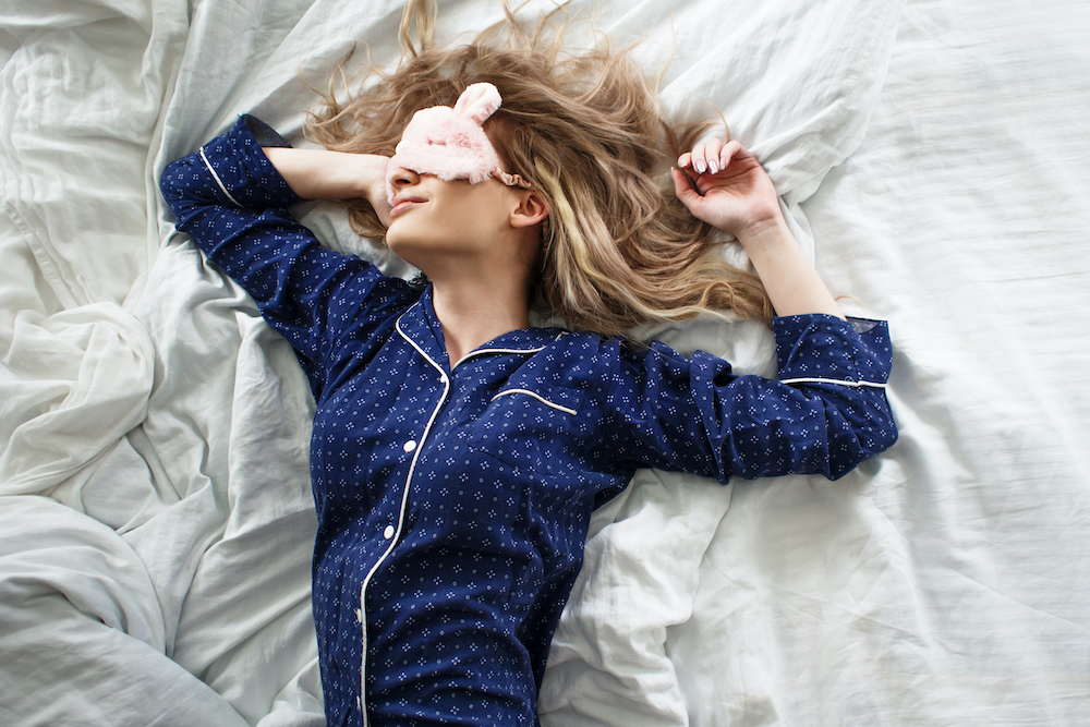 blonde woman laying in bed with eye mask and navy blue pajamas